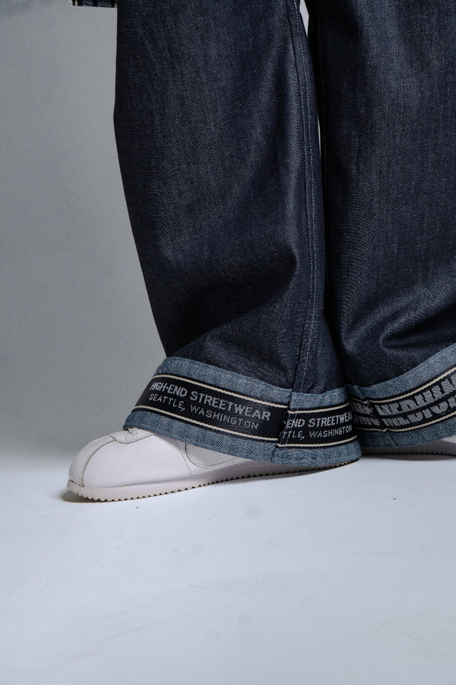 Close-up of dark blue jeans with brand logo on cuffs and white sneakers on a gray background
