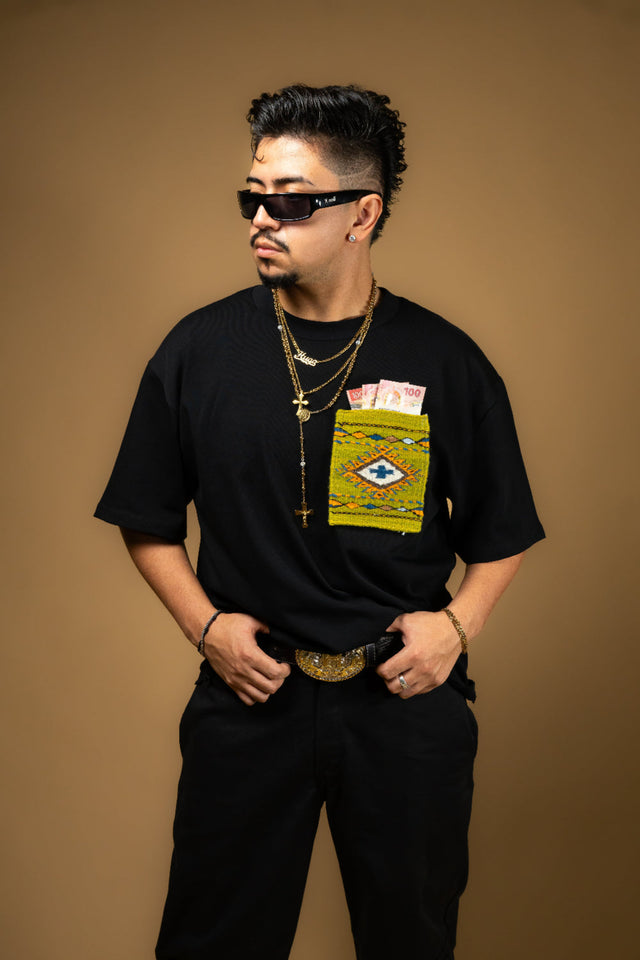 Man wearing a black t-shirt with a Artesanal pocket, sunglasses, and multiple necklaces on a brown background. Oaxaca woven pocket, Indigenous handmade streetwear, Mexican artisan clothing, naturally dyed wool, one of one t-shirt, Seattle streetwear brand, artisanal fashion, handwoven textile apparel, marigold dye, cochineal dye, indigo dye, sheep wool pocket tee.