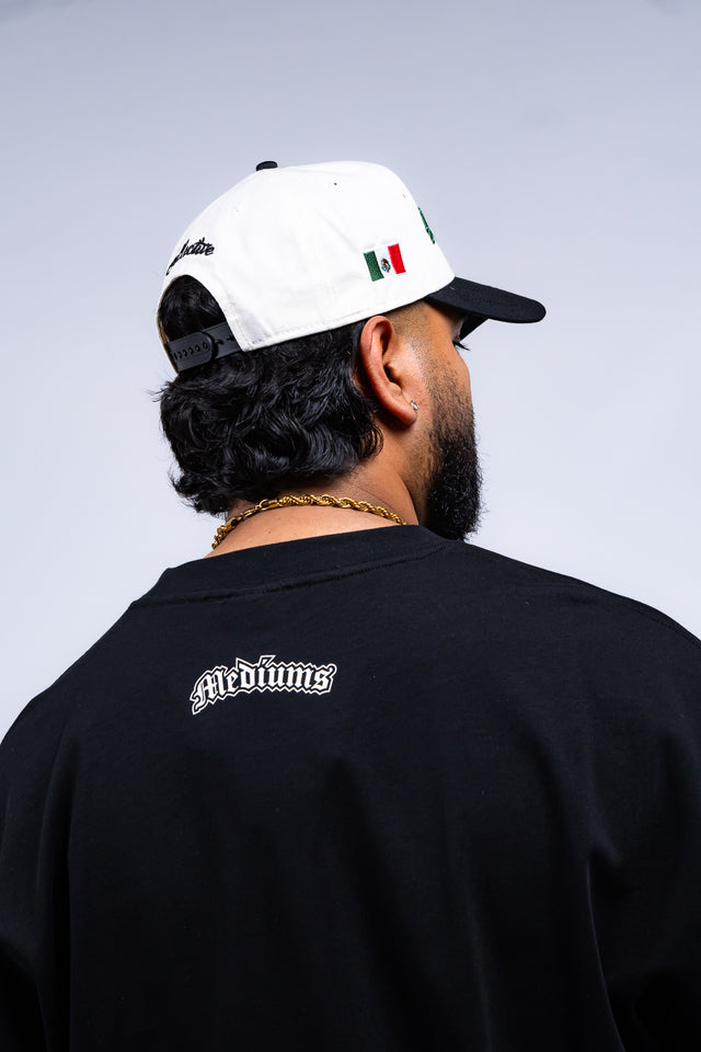 Person wearing a black t-shirt with 'Mediums' logo and a white cap with black visor and Mexican flag design.