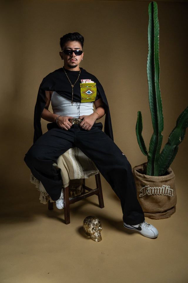 Model sitting on a chair with a cactus plant and skull on a brown background, Man wearing a black t-shirt with a Artesanal pocket, sunglasses, and multiple necklaces on a brown background. Oaxaca woven pocket, Indigenous handmade streetwear, Mexican artisan clothing, naturally dyed wool, one of one t-shirt, Seattle streetwear brand, artisanal fashion, handwoven textile apparel, marigold dye, cochineal dye, indigo dye, sheep wool pocket tee.