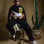 Model sitting on a chair with a cactus plant and skull on a brown background, Man wearing a black t-shirt with a Artesanal pocket, sunglasses, and multiple necklaces on a brown background. Oaxaca woven pocket, Indigenous handmade streetwear, Mexican artisan clothing, naturally dyed wool, one of one t-shirt, Seattle streetwear brand, artisanal fashion, handwoven textile apparel, marigold dye, cochineal dye, indigo dye, sheep wool pocket tee.