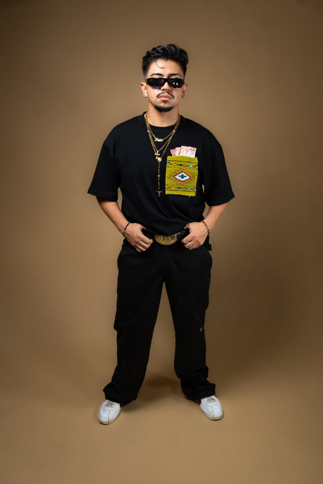 Man wearing a black t-shirt with a Artesanal pocket, sunglasses, and multiple necklaces on a brown background. Oaxaca woven pocket, Indigenous handmade streetwear, Mexican artisan clothing, naturally dyed wool, one of one t-shirt, Seattle streetwear brand, artisanal fashion, handwoven textile apparel, marigold dye, cochineal dye, indigo dye, sheep wool pocket tee.