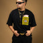 Man wearing a black t-shirt with a Artesanal pocket, sunglasses, and multiple necklaces on a brown background. Oaxaca woven pocket, Indigenous handmade streetwear, Mexican artisan clothing, naturally dyed wool, one of one t-shirt, Seattle streetwear brand, artisanal fashion, handwoven textile apparel, marigold dye, cochineal dye, indigo dye, sheep wool pocket tee.