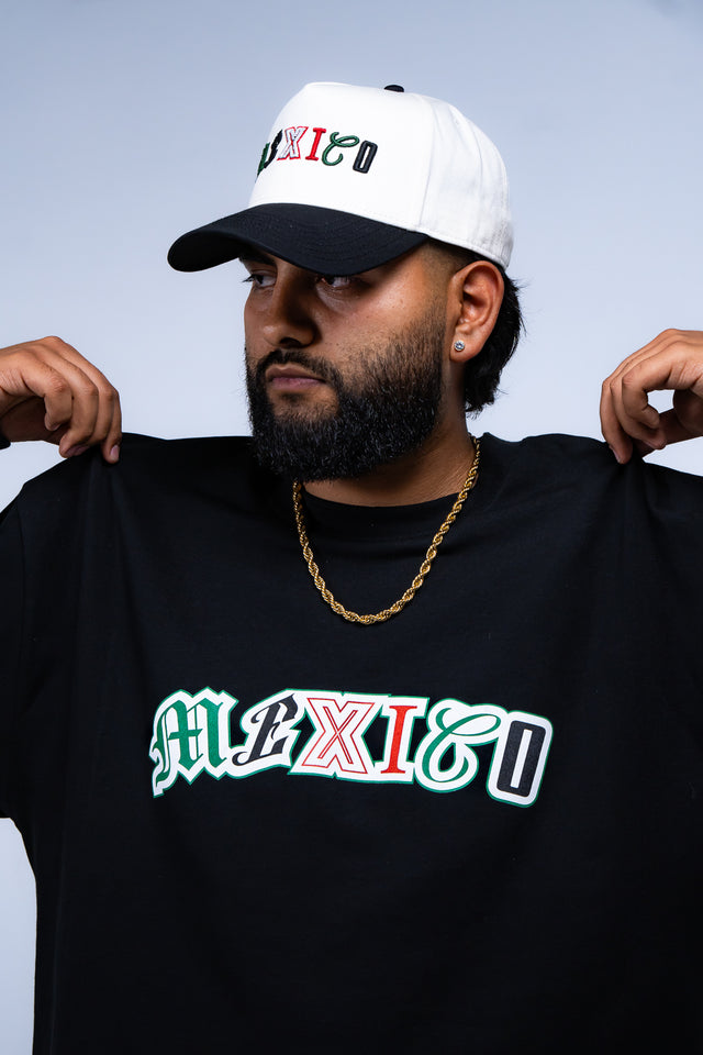 Person wearing a black 'MEXICO' t-shirt and white cap with text on a light blue background Mexico graphic tee, Mexico long sleeve shirt, Mexican streetwear, Latino streetwear, Mediums Collective, Seattle streetwear, Mexico shirt, Mexico soccer shirt, fútbol fashion, Mexican heritage clothing, men’s graphic tee, men’s long sleeve graphic shirt, pine green tee, black long sleeve shirt, white long sleeve shirt, cultural streetwear, World Cup outfit, Mexico capsule, Latino fashion brand, Mexican inspired appare
