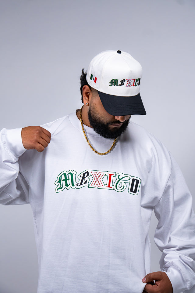 Person wearing a white long-sleeve shirt with 'MEXICO' printed on it, a white cap, and a black cap with visible branding. Mexico graphic tee, Mexico long sleeve shirt, Mexican streetwear, Latino streetwear, Mediums Collective, Seattle streetwear, Mexico shirt, Mexico soccer shirt, fútbol fashion, Mexican heritage clothing, men’s graphic tee, men’s long sleeve graphic shirt, pine green tee, black long sleeve shirt, white long sleeve shirt, cultural streetwear, World Cup outfit, Mexico capsule, Latino fashion